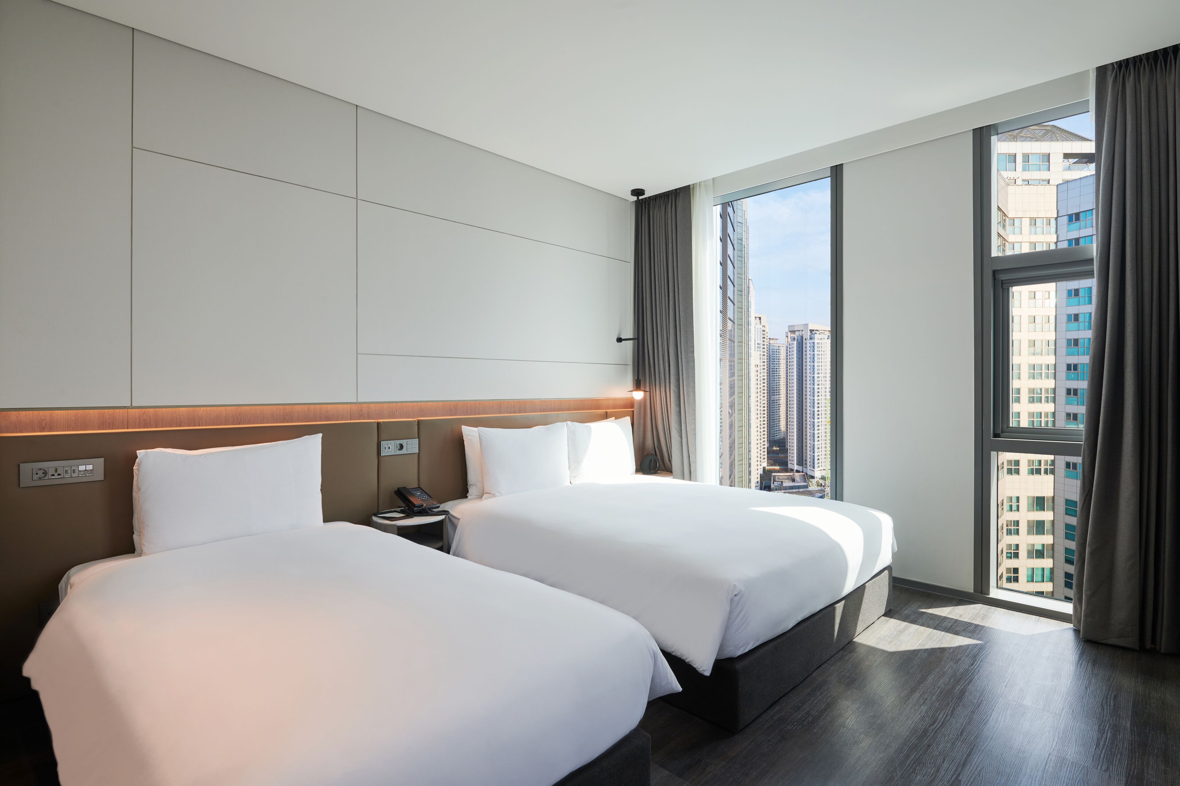 Habitación de hotel con dos camas, una mesita de noche, un teléfono y un ventanal grande con cortinas.