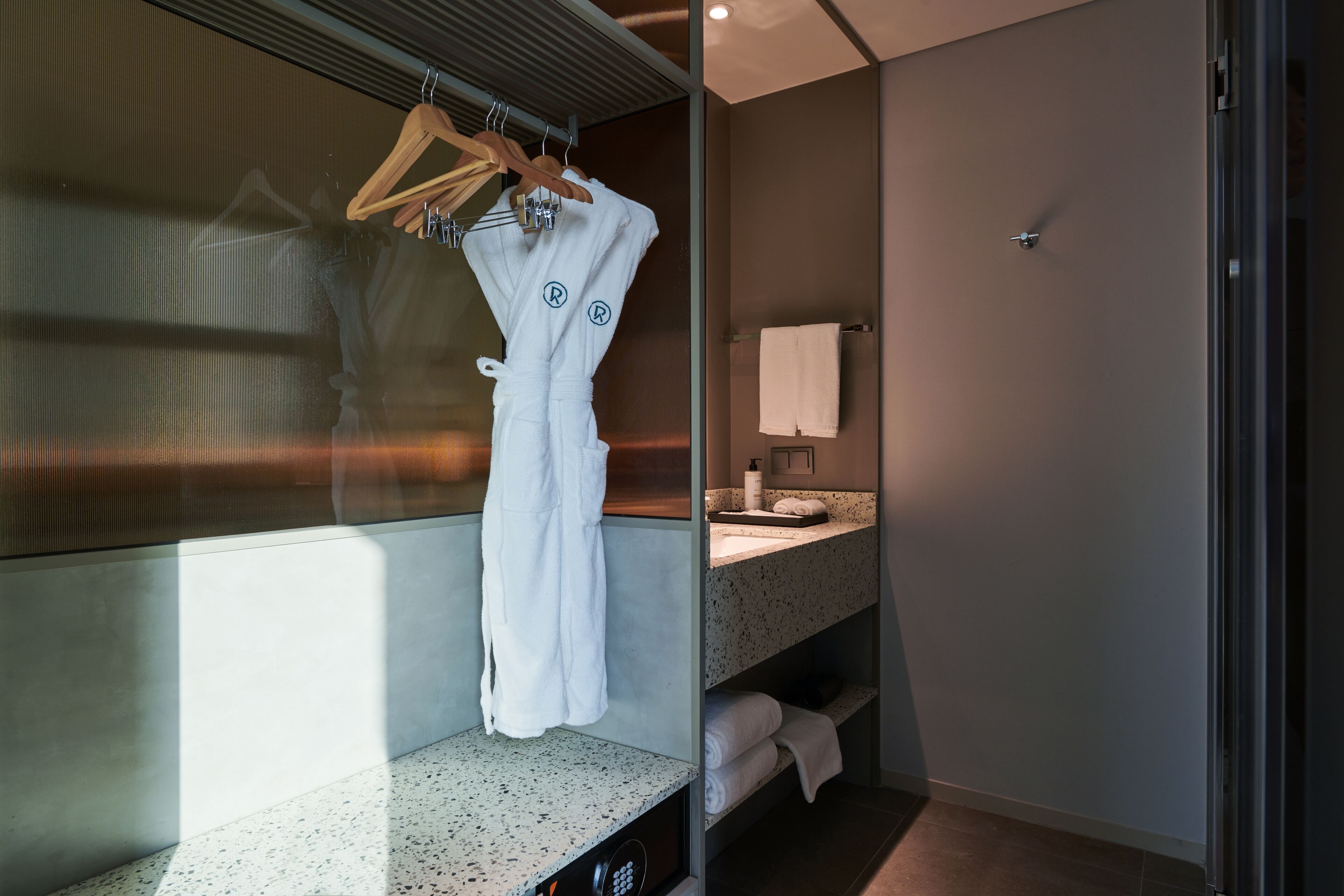 A modern bathroom with a glass shower enclosure, a towel rack with a white robe, a countertop with a soap dispenser, and a mirror.