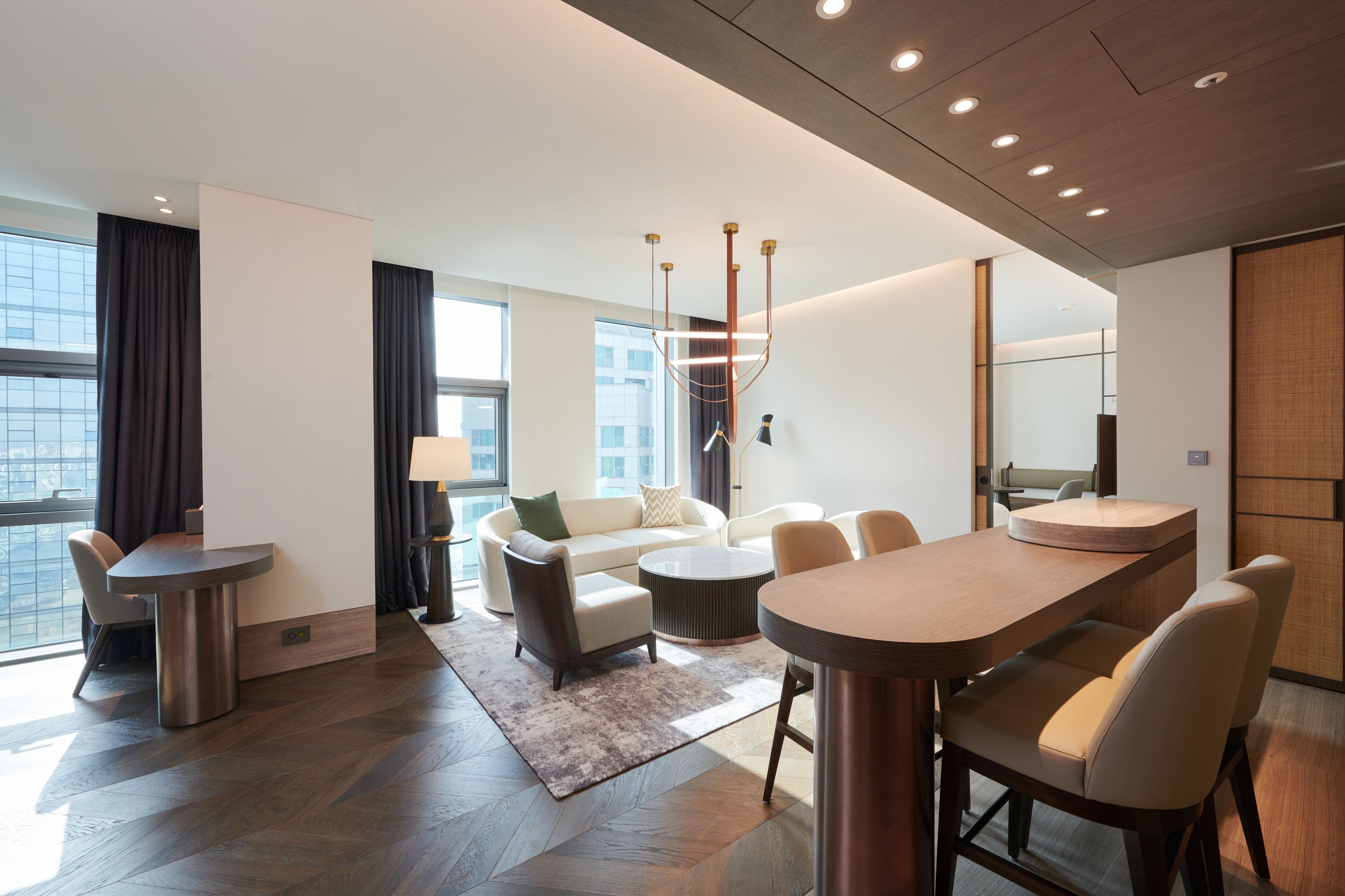 Une chambre d’hôtel moderne dotée d’un coin repas, d’un ensemble de canapés et de grandes fenêtres offrant une vue sur la ville.