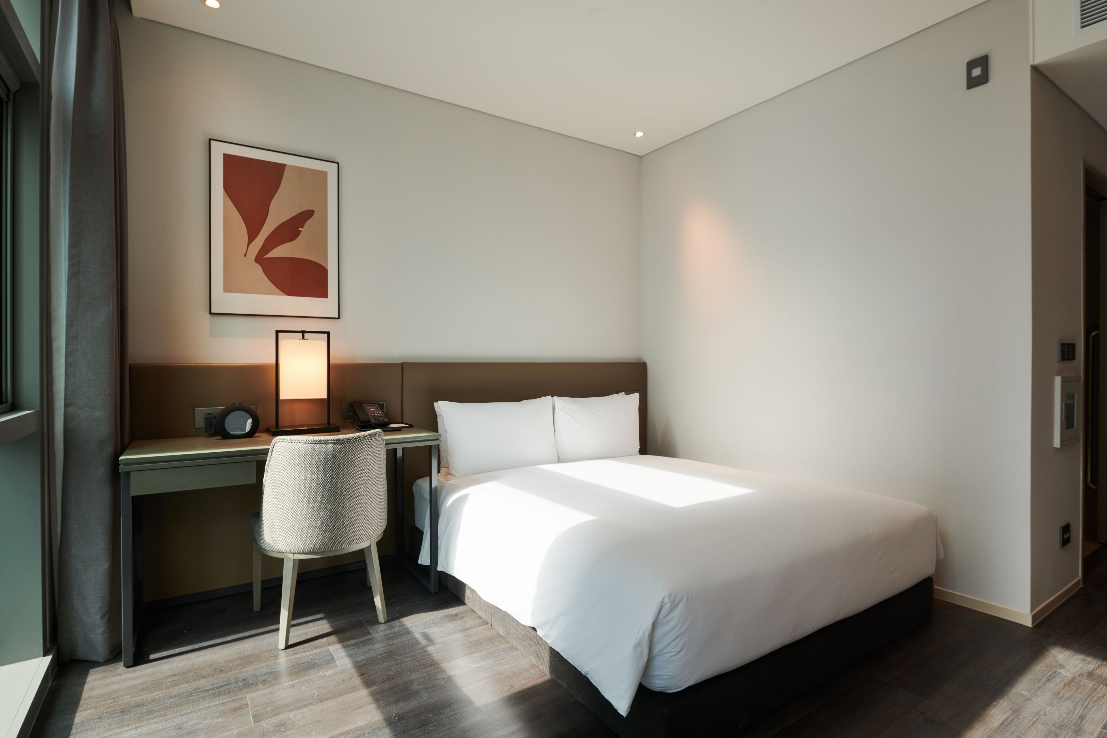 A hotel room with a large bed, a desk with a chair, a lamp, and a wall-mounted artwork.