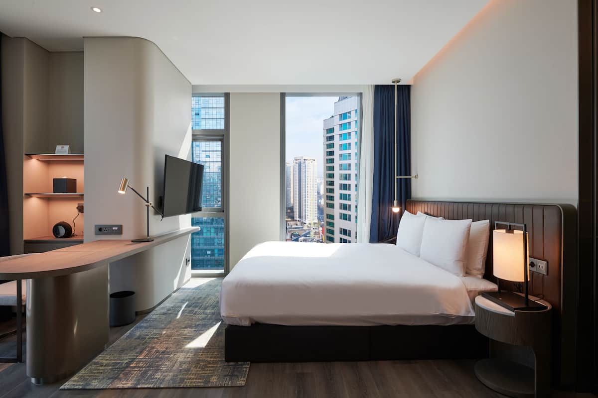 A modern hotel room with a large bed, a desk with a flat-screen TV, a bedside table with a lamp, and a view of the cityscape through large windows.