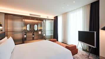 A modern hotel room with a large bed, a TV, a walk-in closet, and a bathroom with a mirror and vanity.