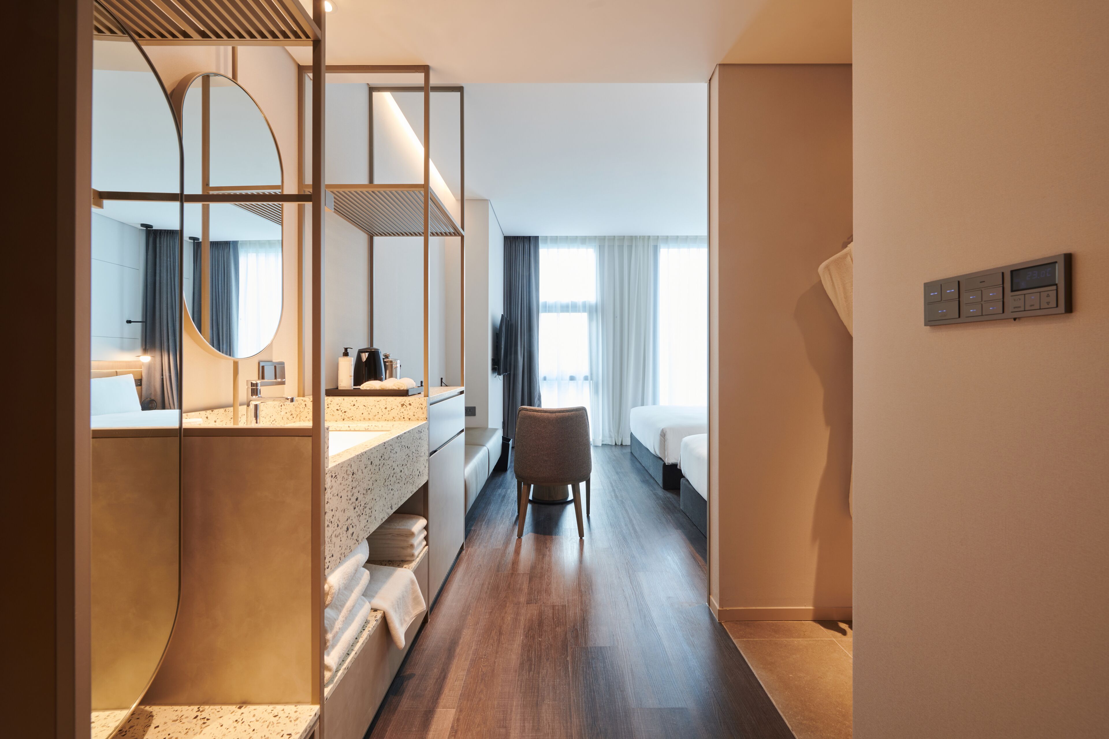 Quarto de hotel moderno com piso de madeira, espelho grande, banheiro com pia e amenities, cama e painel de controle na parede.