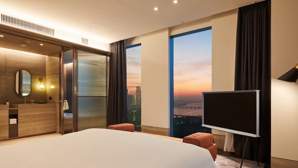 A modern hotel room with a large bed, a TV, a city view, and a walk-in closet.