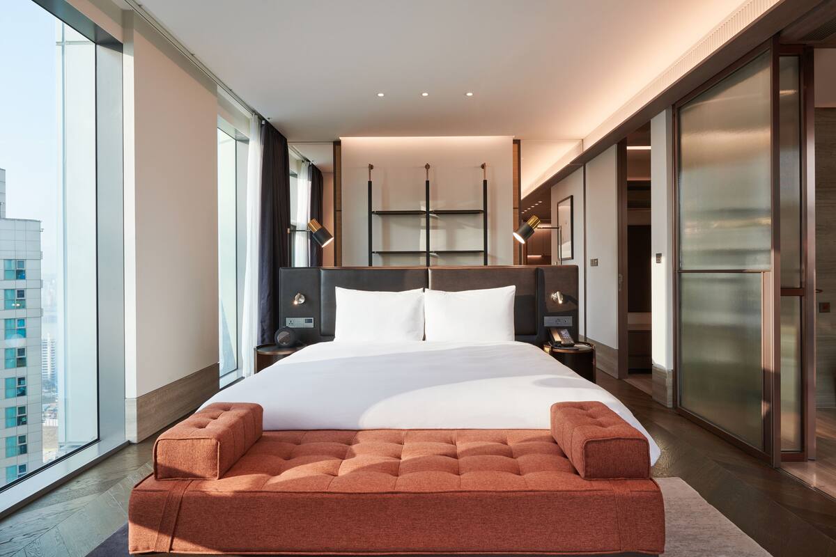 A modern hotel room with a large bed, a seating area, a nightstand, and a view of the city.