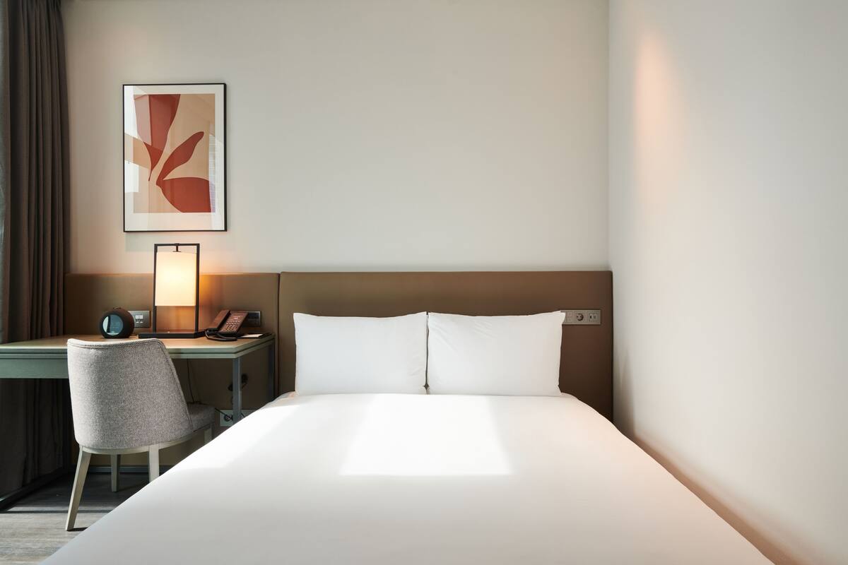 A hotel room with a bed, a desk, a chair, a lamp, and a framed picture on the wall.