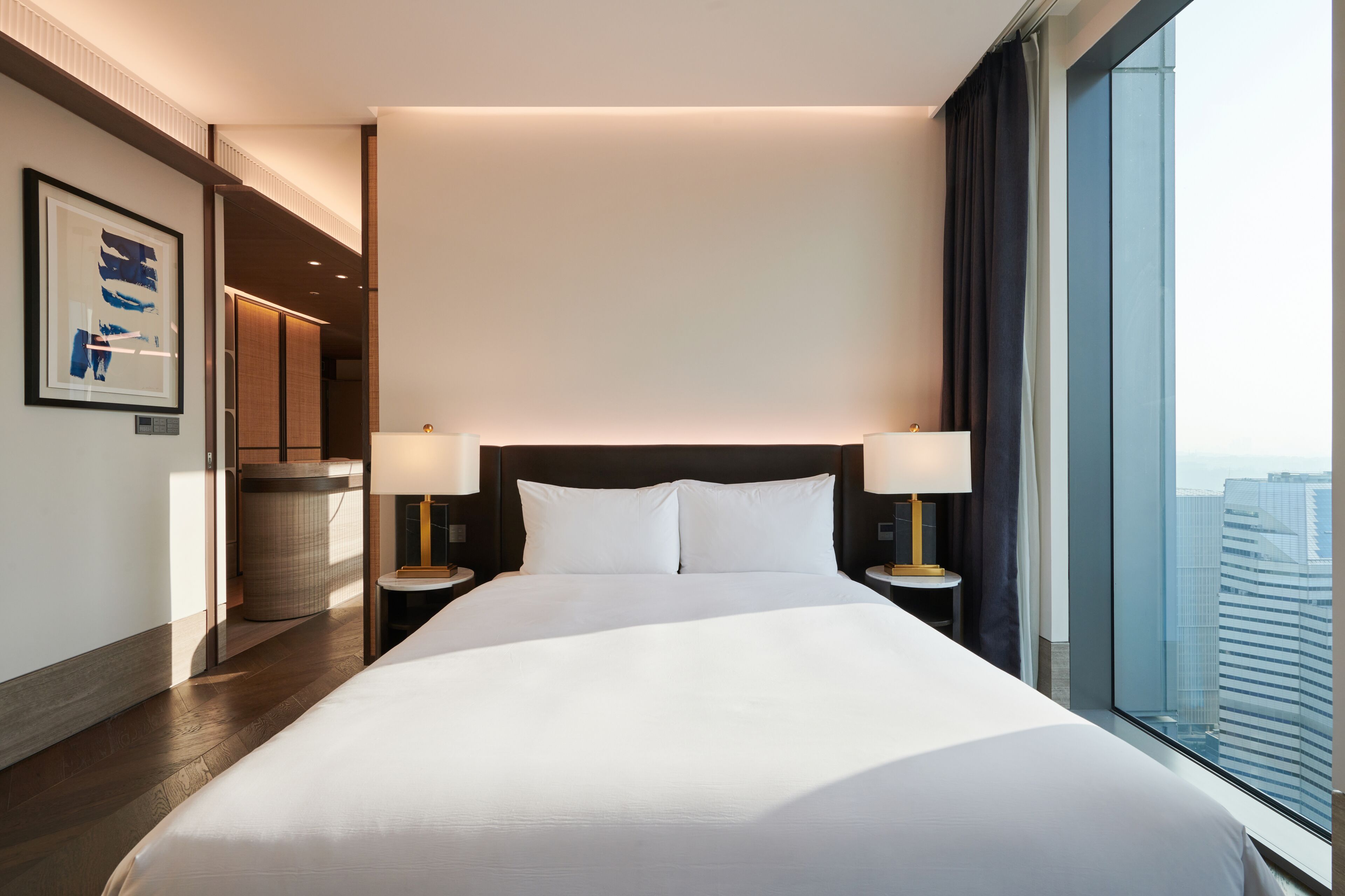 A modern hotel room with a large bed, two bedside tables with lamps, a large window with a city view, and a framed artwork on the wall.
