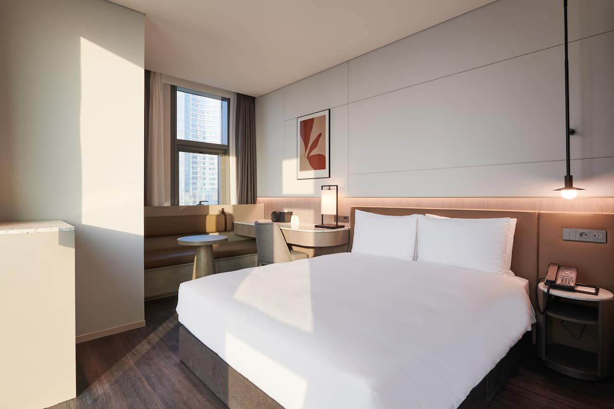 A hotel room with a large bed, a desk with a chair, a small table, a sofa, a window with curtains, a wall-mounted phone, and a light fixture.
