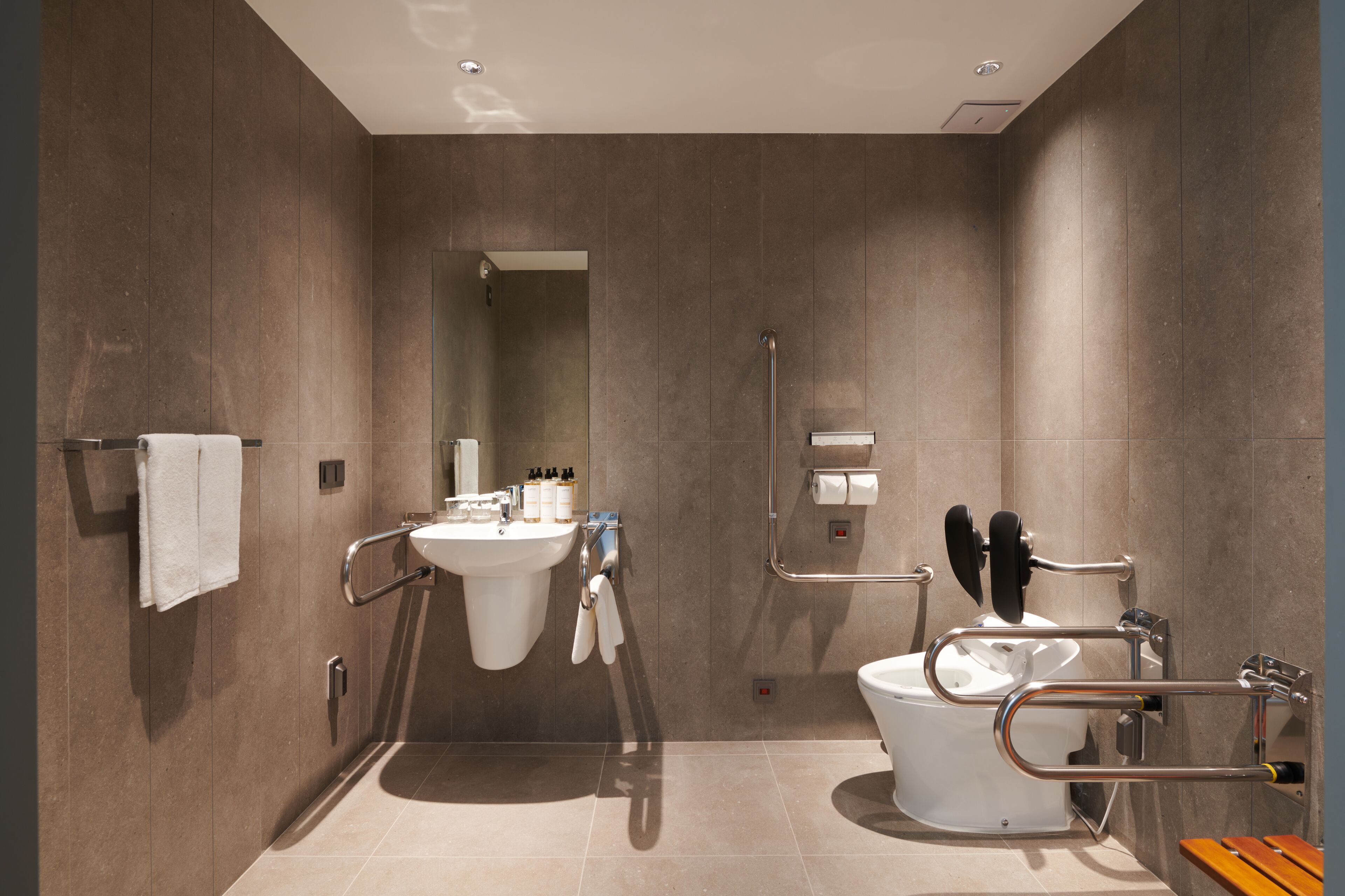 Un baño moderno con lavamanos, inodoro y ducha. Cuenta con barras de apoyo y un banco.