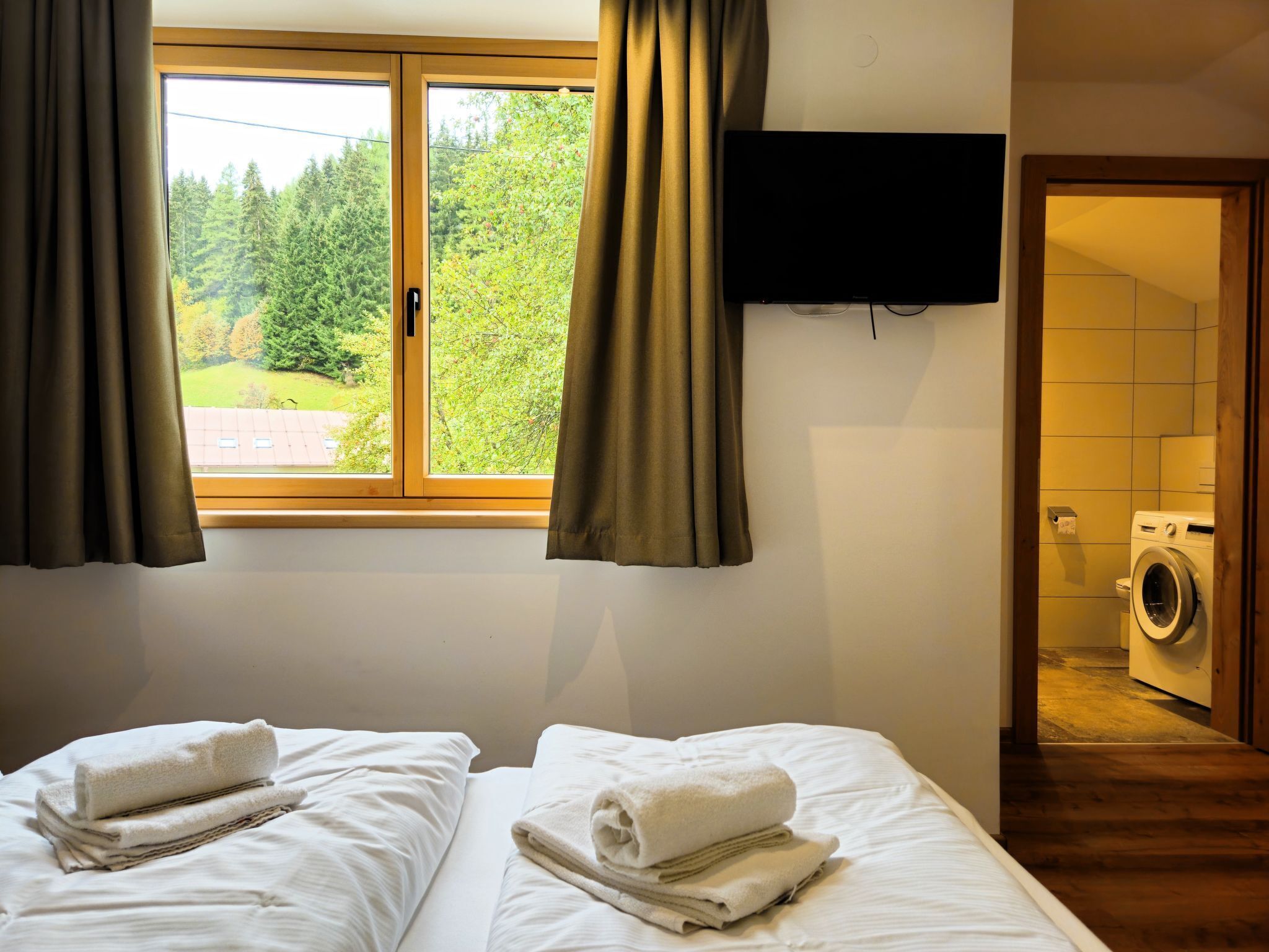 A hotel room with two beds, a window with curtains, a flat-screen TV mounted on the wall, and a washing machine in the corner.