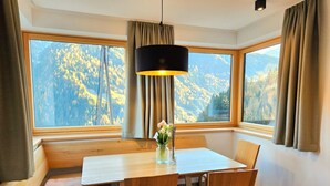 A dining area with a wooden table and chairs, a vase with flowers, and large windows with a mountain view.