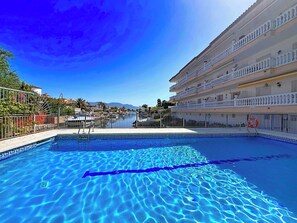 A swimming pool with clear blue water, surrounded by a white building with balconies, and a clear sky.
