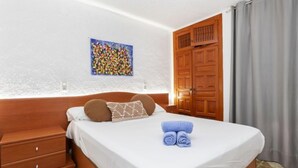 Ein ordentlich bezogenes Bett mit weißer Bettdecke und zwei Kissen, ein Nachttisch aus Holz mit einer Schublade, ein Holzschrank und ein farbenfrohes abstraktes Gemälde an der Wand.