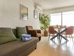 A living room with a sofa, armchairs, a dining table with chairs, a potted plant, and framed pictures on the wall.
