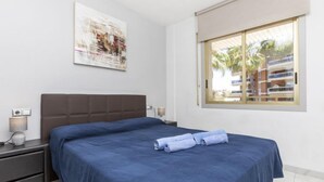 Camera da letto con un letto grande, un comodino, una lampada e una finestra vista edifici e palme.