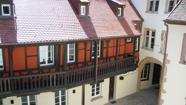 Ein traditionelles europäisches Gebäude mit rotem Ziegeldach, einem Balkon mit schwarzen Geländern und einem Kopfsteinpflasterhof.
