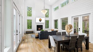 A spacious living room with a fireplace, a dining area with a dark wooden table and wicker chairs, and large windows offering views of the surrounding forest.
