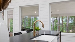 A modern kitchen with a black countertop, a gold faucet, and a view of the forest through large windows.