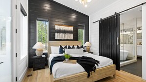 A modern bedroom with a wooden bed, two bedside tables, a large window, and a sliding glass door leading to a bathroom.
