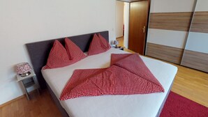 Een netjes opgemaakt bed met een wit-rood geruit dekbed, een grijs hoofdbord en een klein nachtkastje met een lamp.
