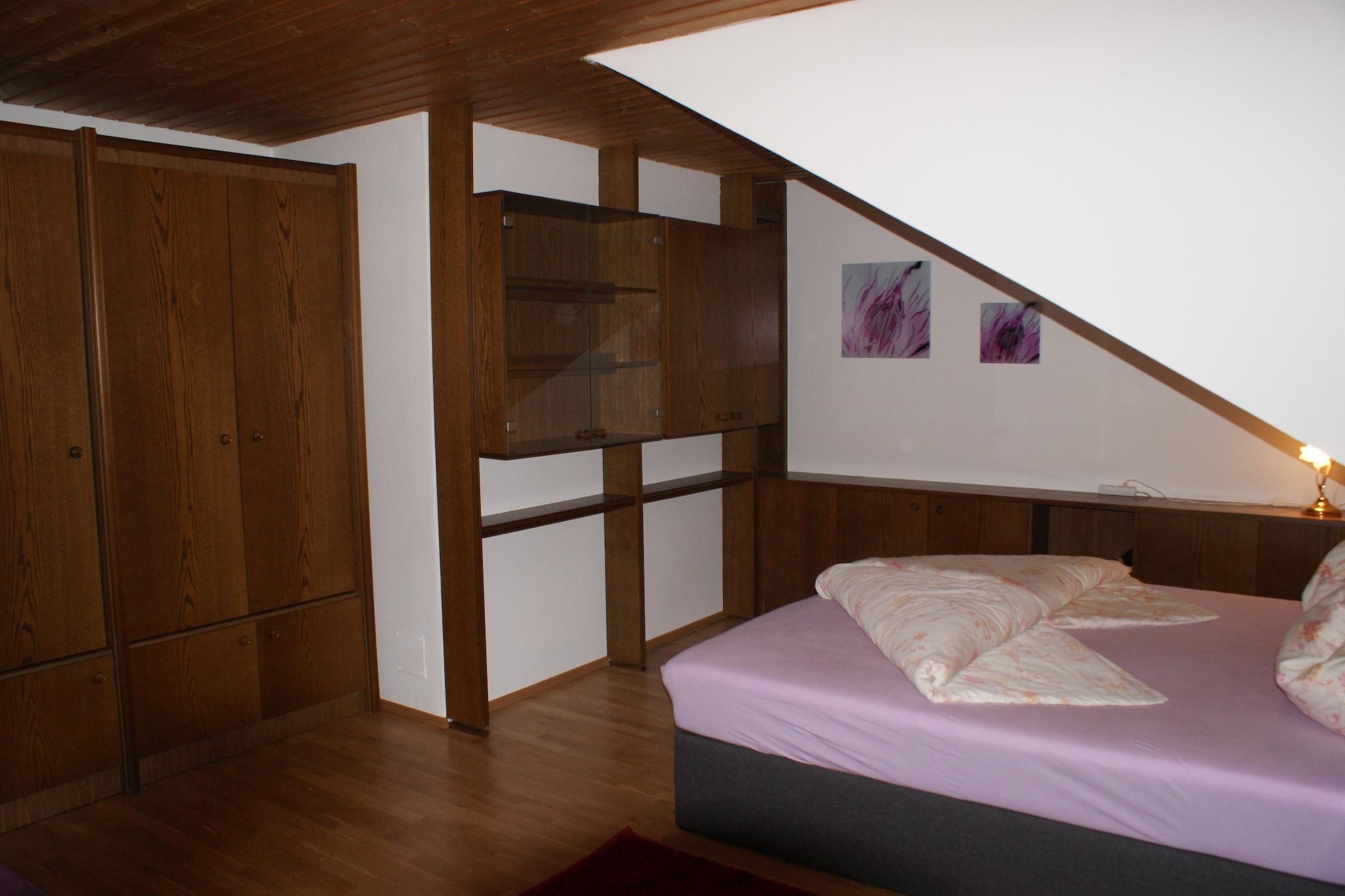 Une chambre avec un plafond en pente, une armoire en bois, un lit recouvert d’une couvre-lit rose et deux tableaux accrochés au mur.