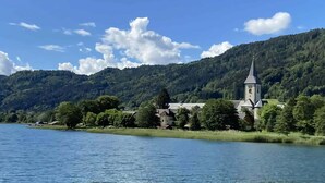 A lakeside view with a church and surrounding greenery. - Holiday Home Bodensdorf near Gerlitzen Ski Area (Bodensdorf am Ossiachersee)