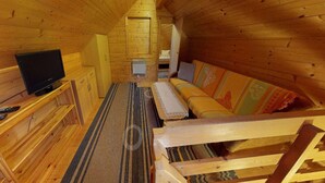 Ein gemütliches Schlafzimmer im Dachgeschoss mit Holzwänden und schrägem Dach. Es beinhaltet einen Fernseher auf einem Holzboden, eine Couch mit bunten Kissen und einen kleinen Tisch mit einer Bank.