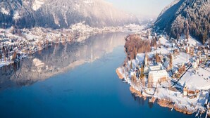 A winter scene with a river, snow-covered buildings, and mountains in the background.