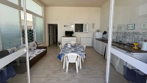 Eine moderne Küche mit Esstisch, weißen Stühlen und einer Couch. An der Wand befindet sich ein Fernseher und ein Fenster mit Jalousien.
