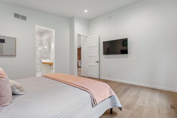 A bedroom with a bed, a TV mounted on the wall, and a bathroom visible through an open door. - Private Riverside Retreat in Saugatuck, MI - 4 Bedrooms, Hot Tub, sleeps 8. (Saugatuck)