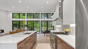 A modern kitchen with a large island, wooden cabinets, and a view of the forest through large windows.