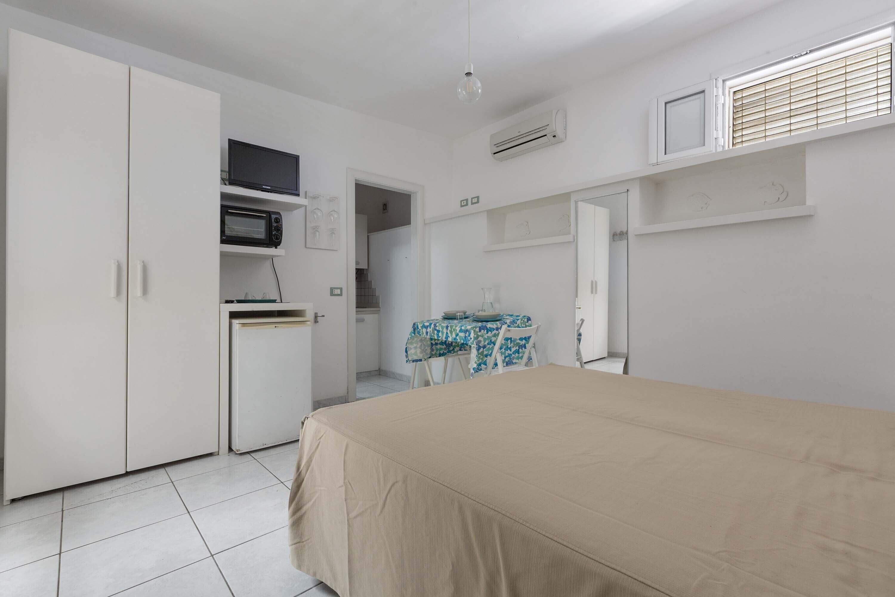A bedroom with a bed, a wardrobe, a small kitchenette, and a dining area.