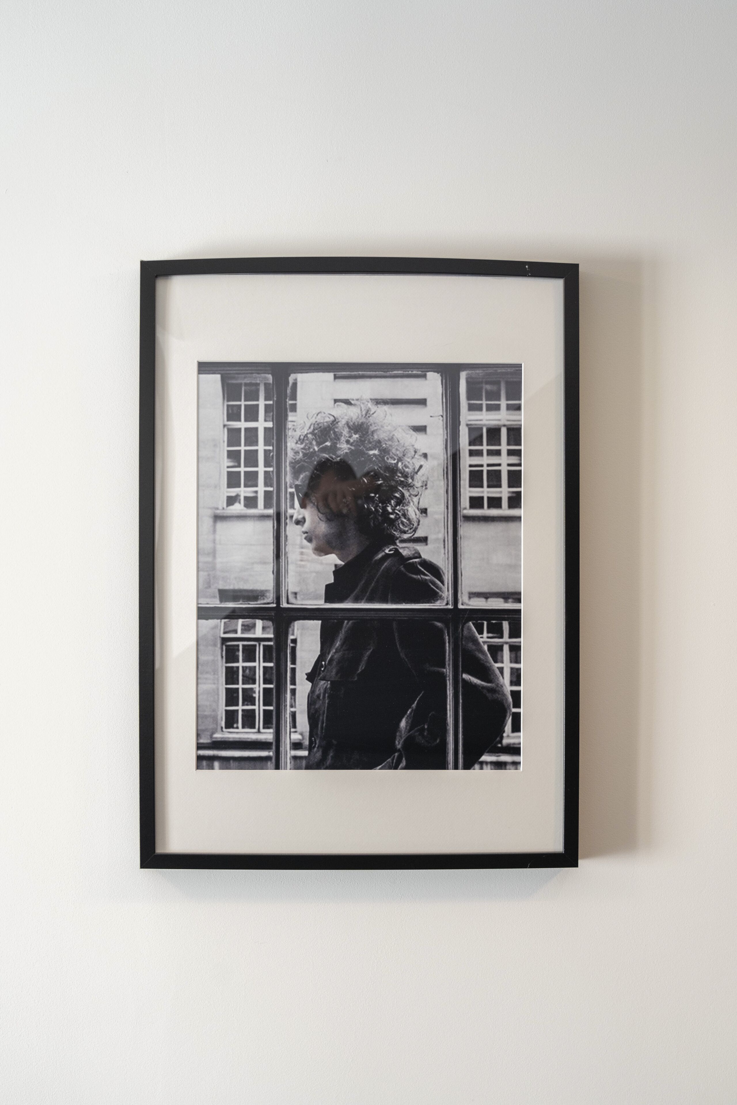 Une photographie en noir et blanc, encadrée, représentant une personne debout devant une fenêtre.