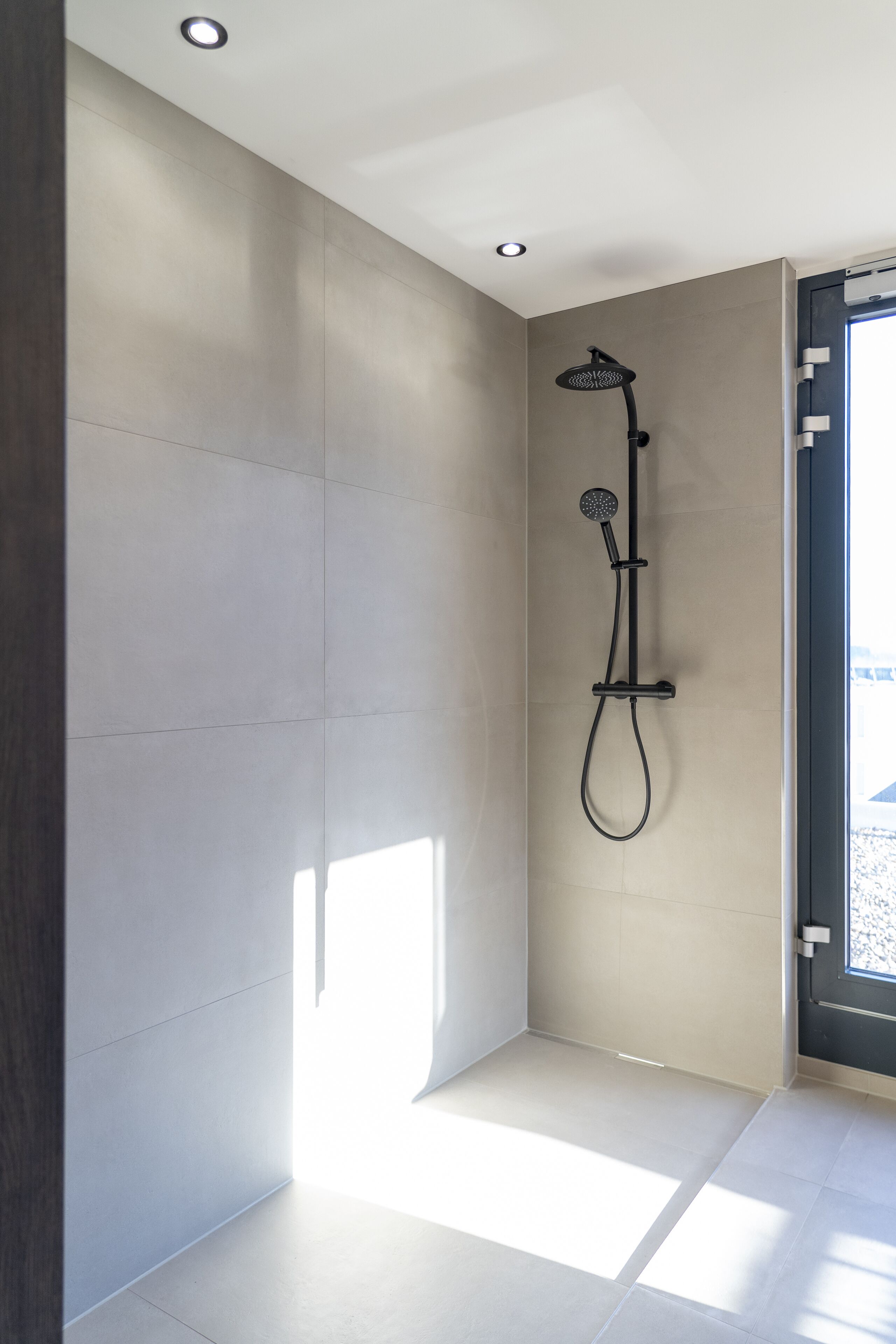 Eine moderne Dusche mit Duschbrause und Reinigungsbürste, ein großes Fenster mit Aussicht und eine Tür mit Griff.