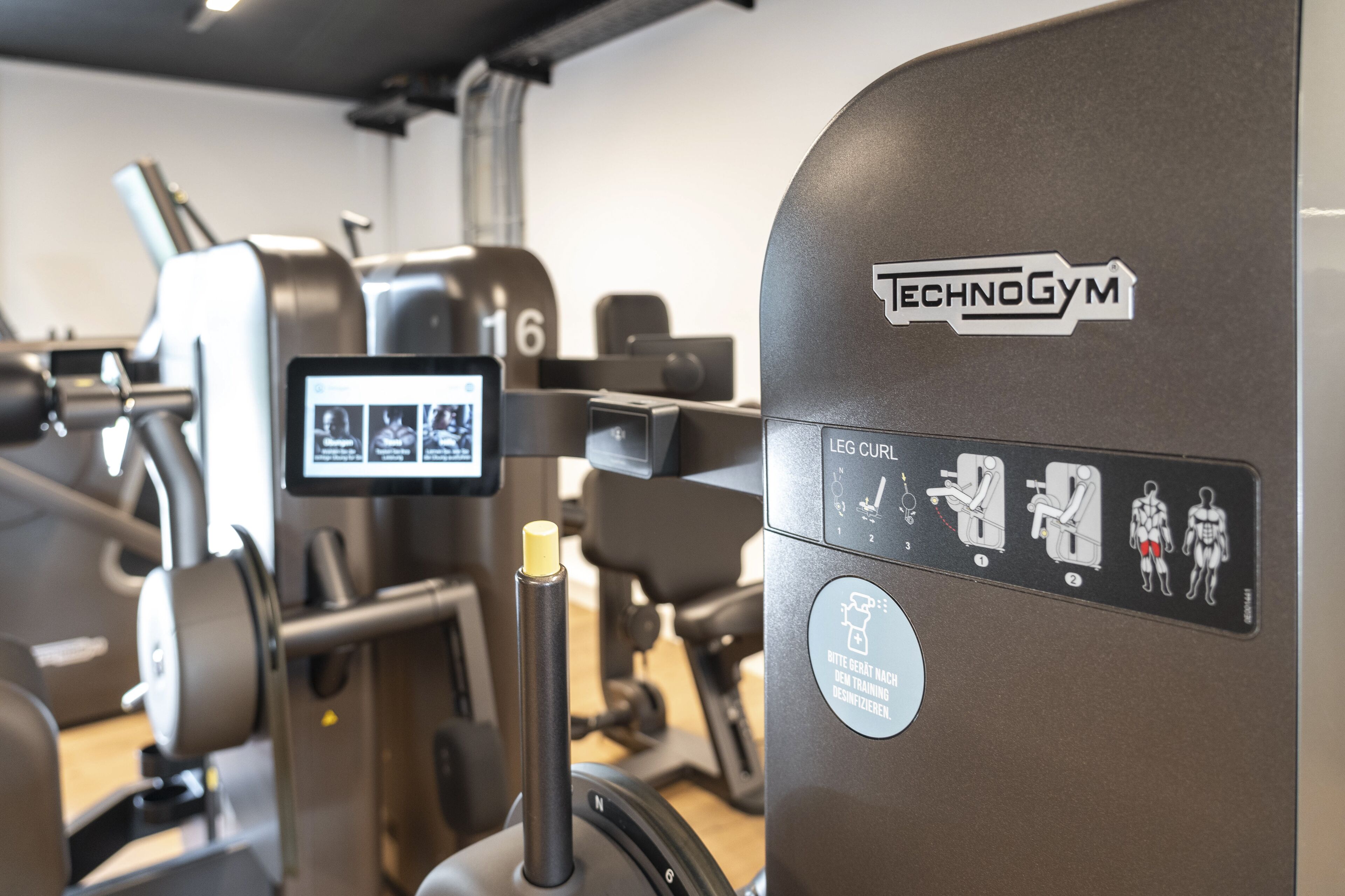 Un gimnasio con diversas máquinas de ejercicio, incluyendo una máquina para curl de piernas con la etiqueta 'TECHNOGYM'.