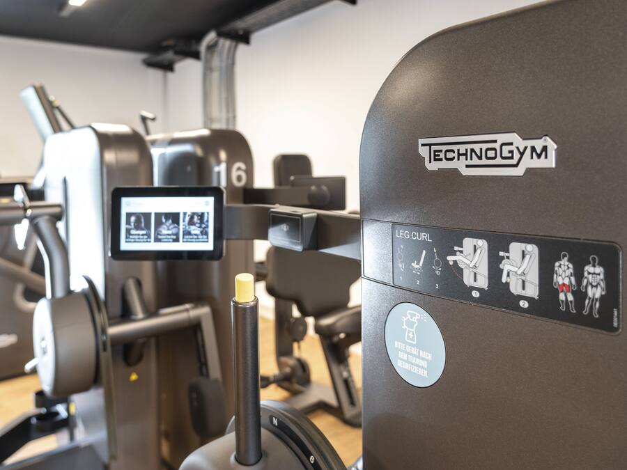 Un gimnasio con diversas máquinas de ejercicio, incluyendo una máquina para curl de piernas con la etiqueta 'TECHNOGYM'.