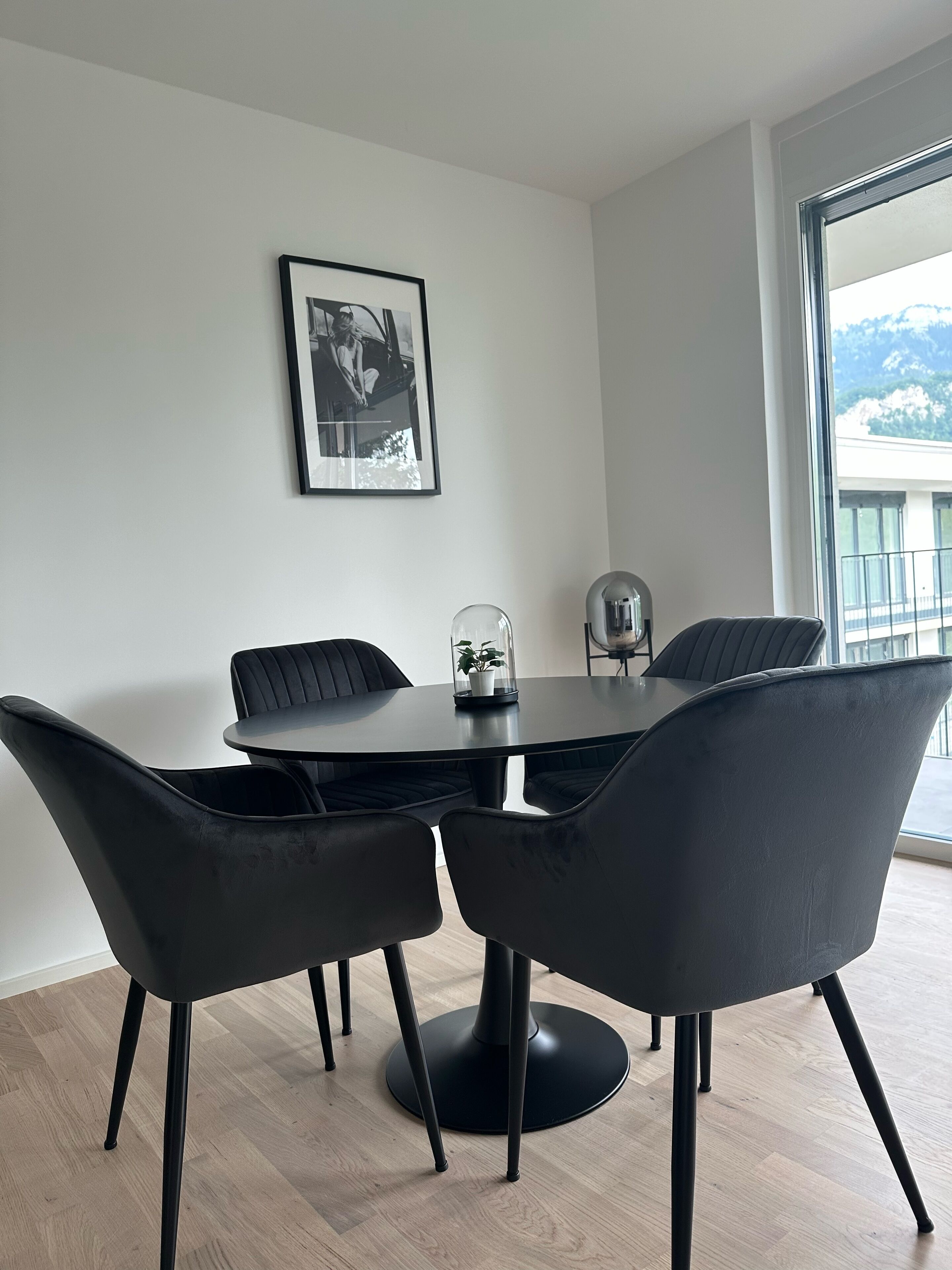 Ein moderner Essbereich mit einem runden Tisch und vier schwarzen Stühlen. An der Wand hängt ein gerahmtes Foto, und auf dem Tisch steht eine Topfpflanze.
