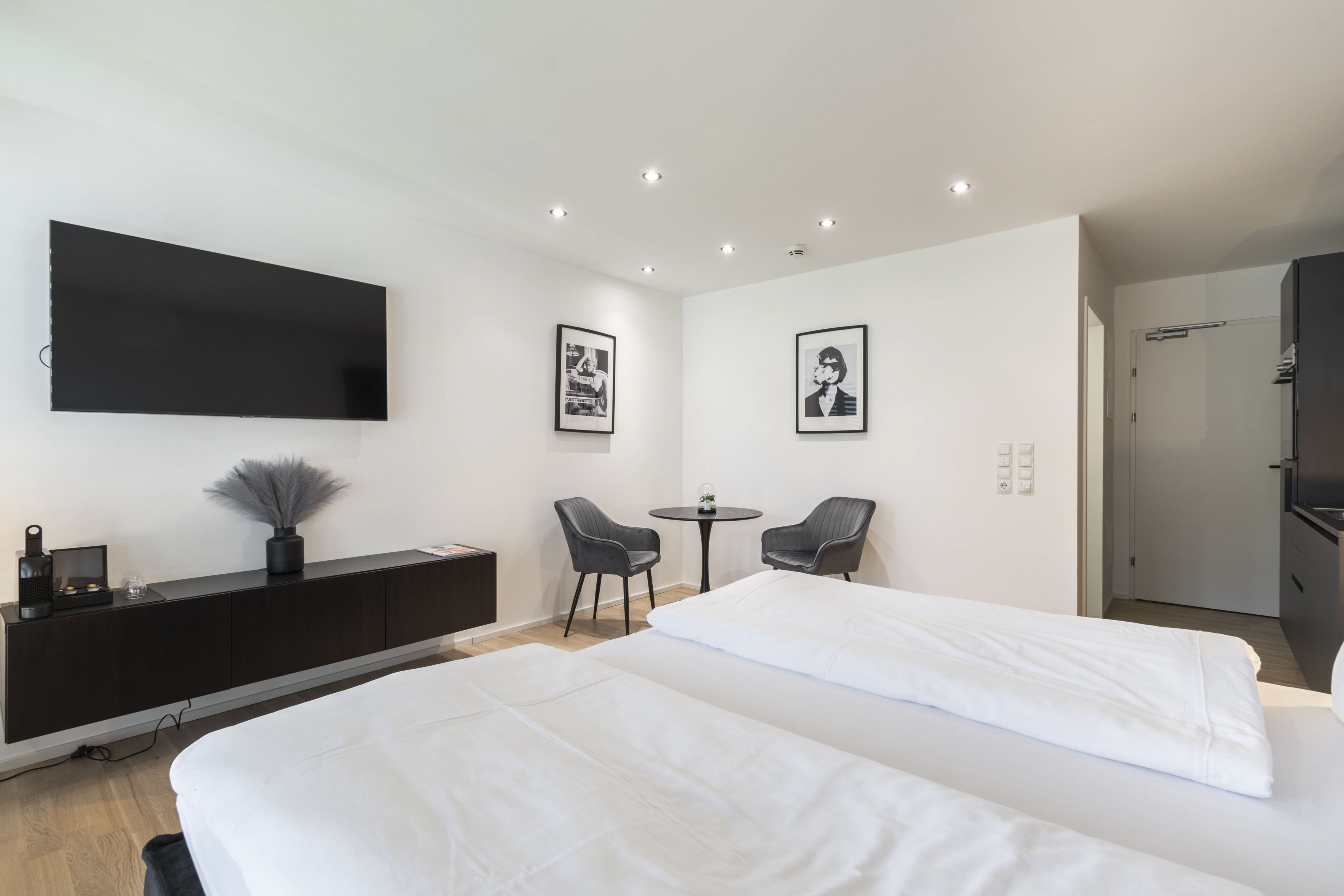 A modern hotel room with a large bed, a TV mounted on the wall, a small table, two chairs, and a dark-colored wardrobe.