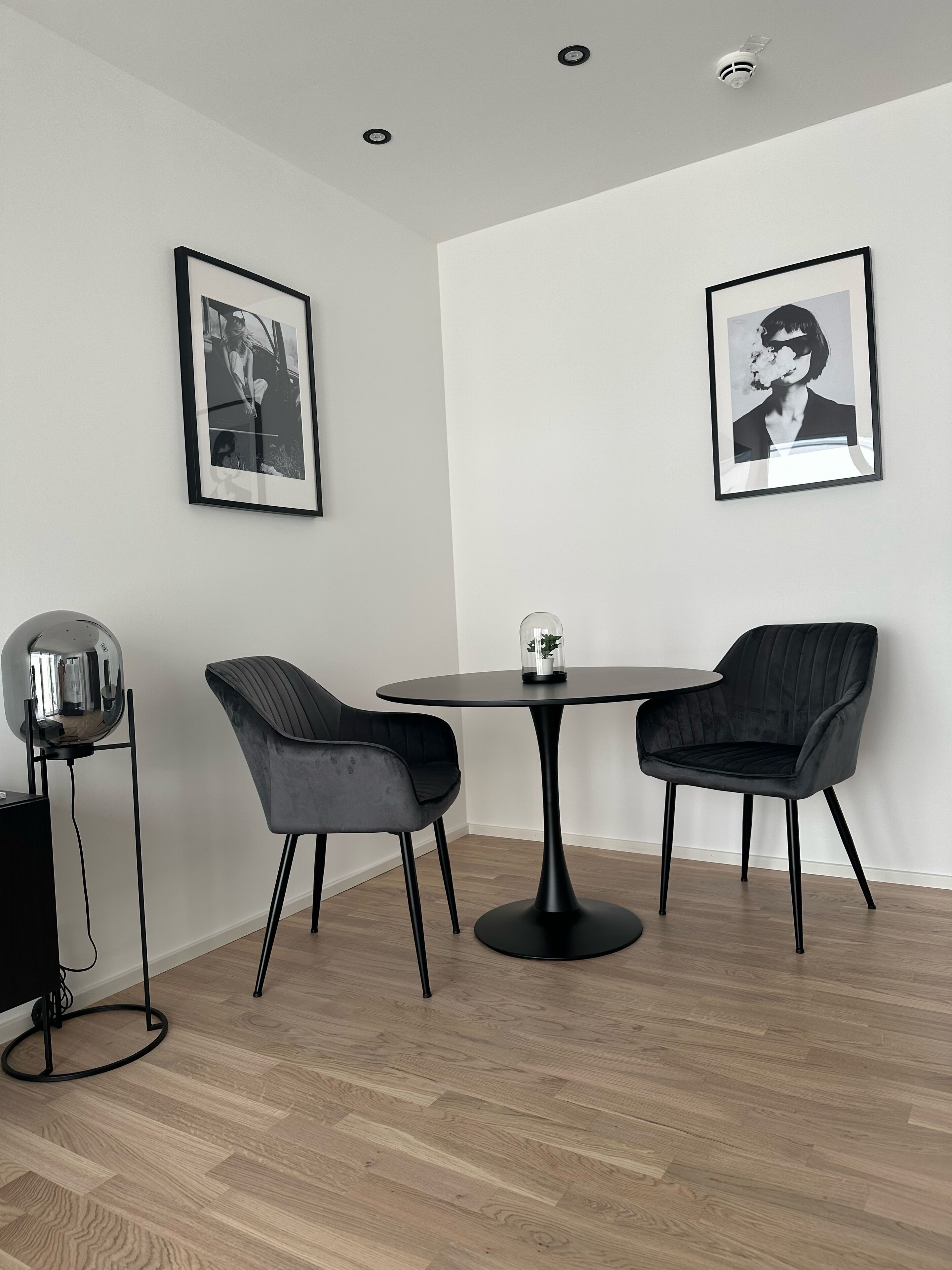 Habitació moderna amb una taula rodona i dues cadires, dues fotografies emmarcades a la paret, una làmpada de peu i una planta petita sobre la taula.