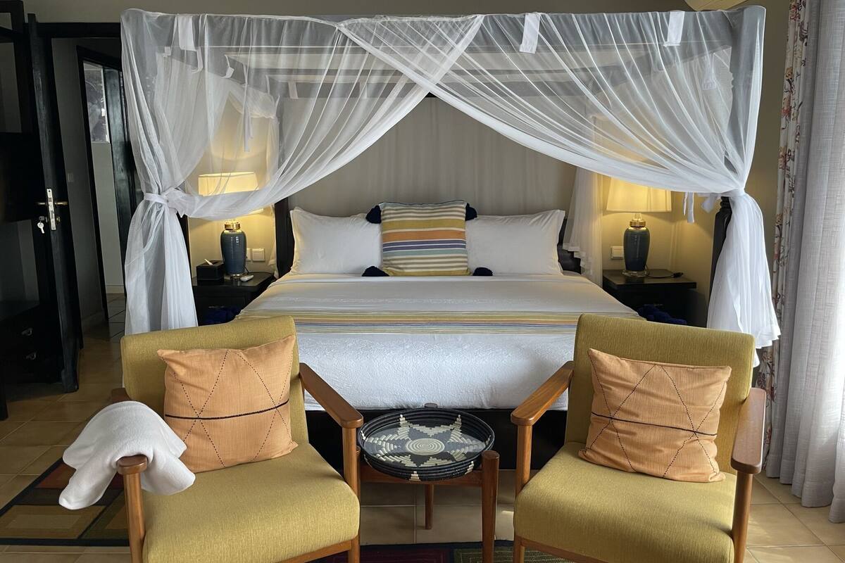 A hotel room with a bed, two armchairs, a nightstand, a lamp, and a mosquito net.