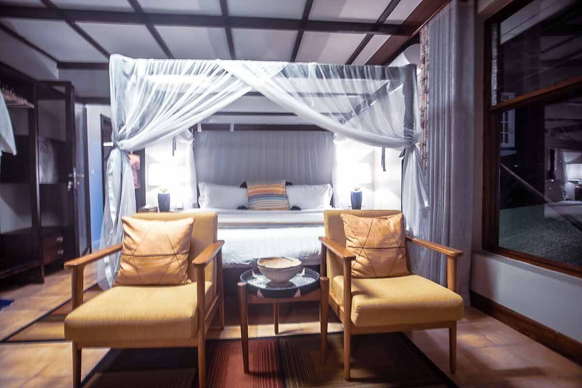 A bedroom with a canopy bed, two armchairs, a bedside table with a bowl, and a window with curtains.