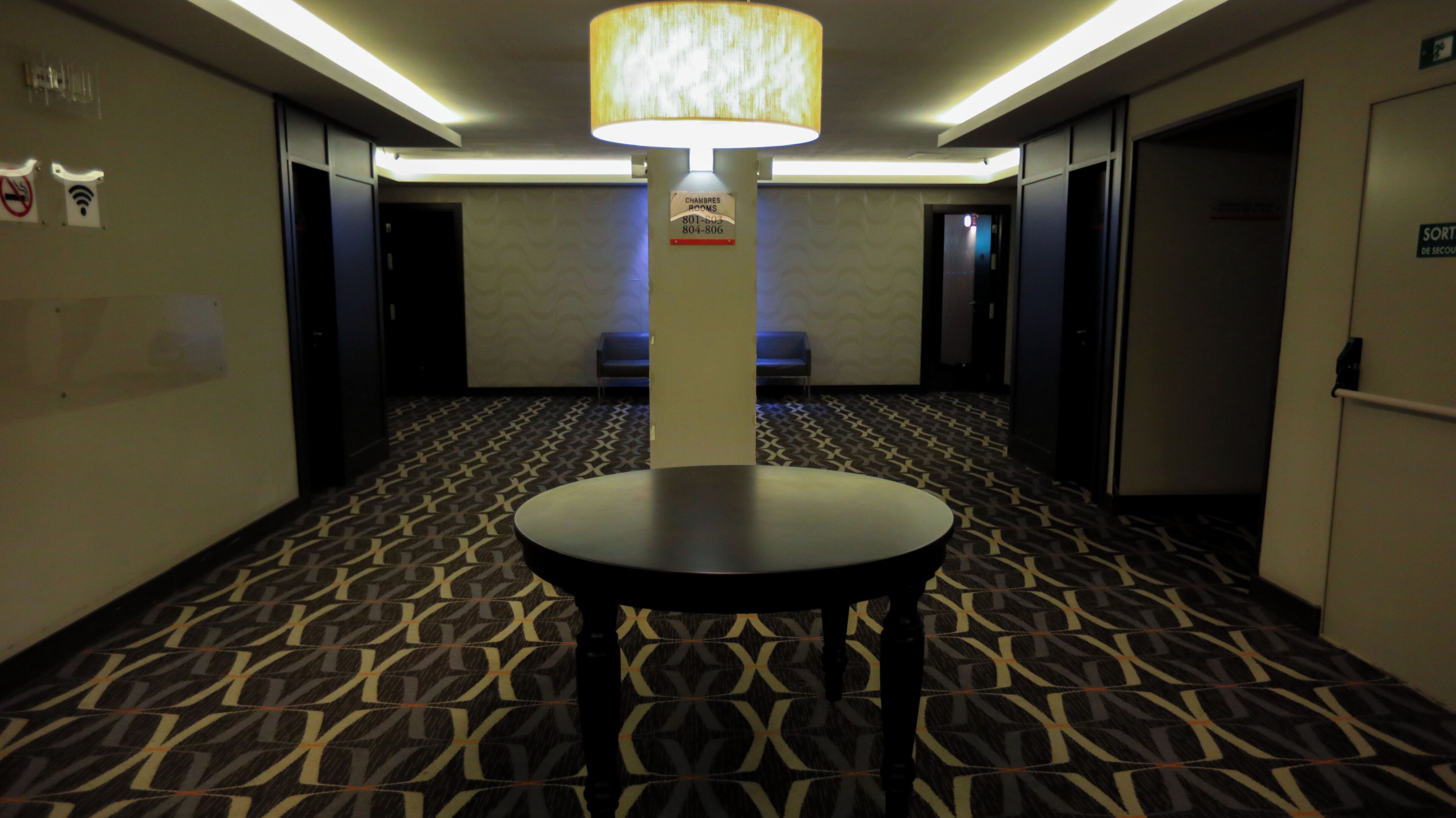 Un couloir d’hôtel avec un tapis à motifs, une table ronde, un pilier portant une enseigne et des portes donnant accès aux chambres.