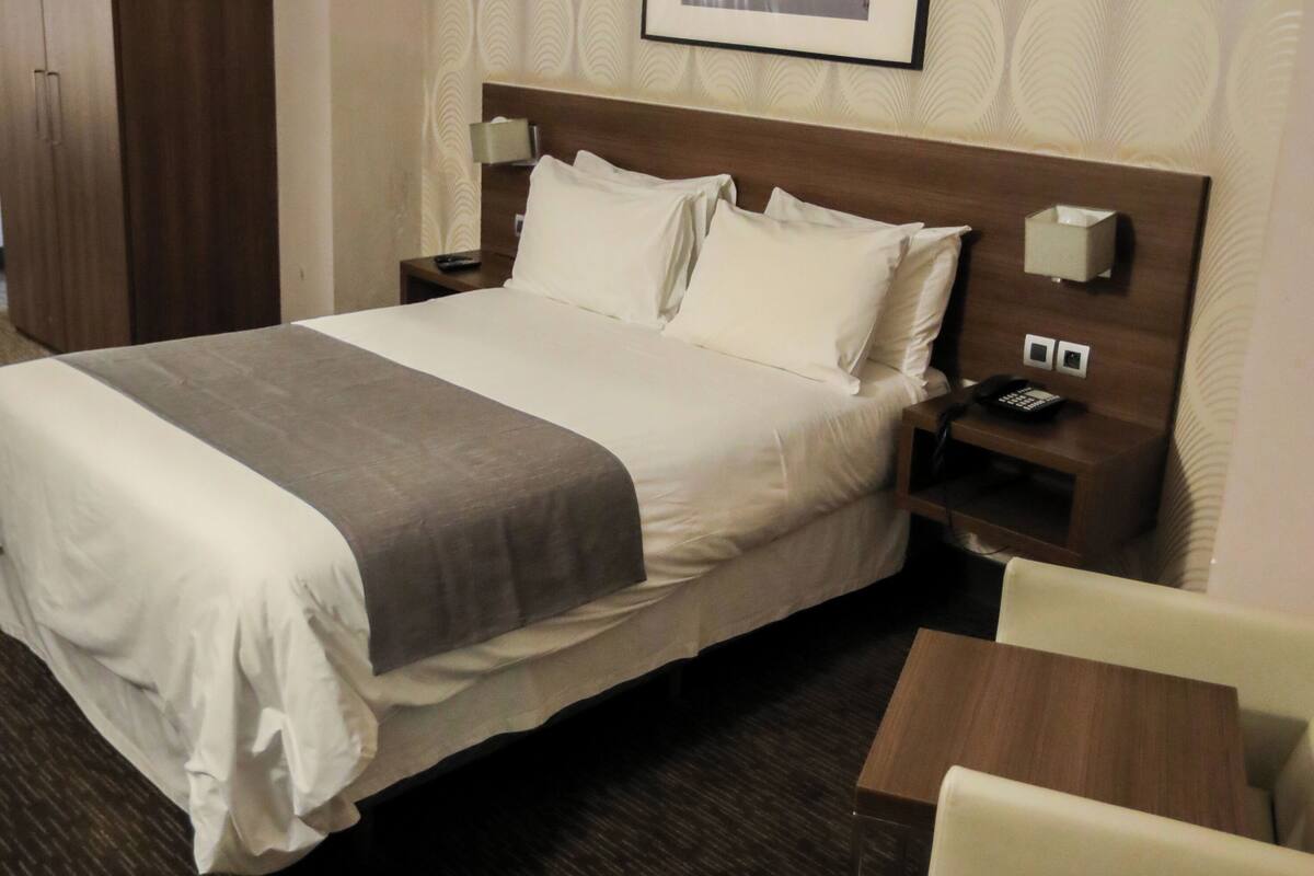 Una habitación de hotel con cama, mesita de noche, silla y un cuadro en la pared.
