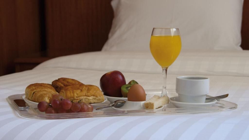 A breakfast tray with croissants, an apple, grapes, a glass of orange juice, a cup of coffee, and a small plate with a pastry on a bed.