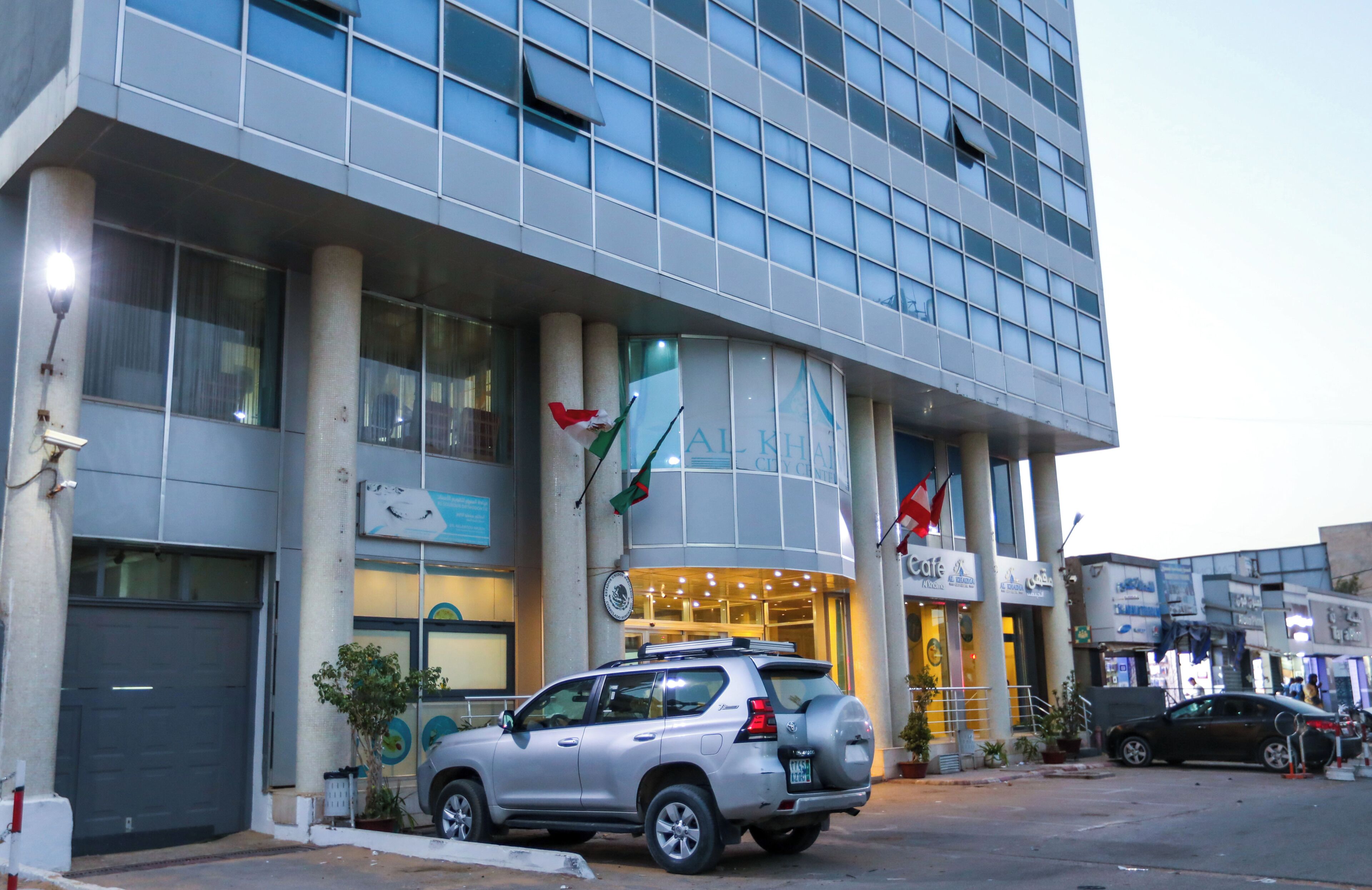 A modern building with a large glass facade, a flag, and a parked SUV.