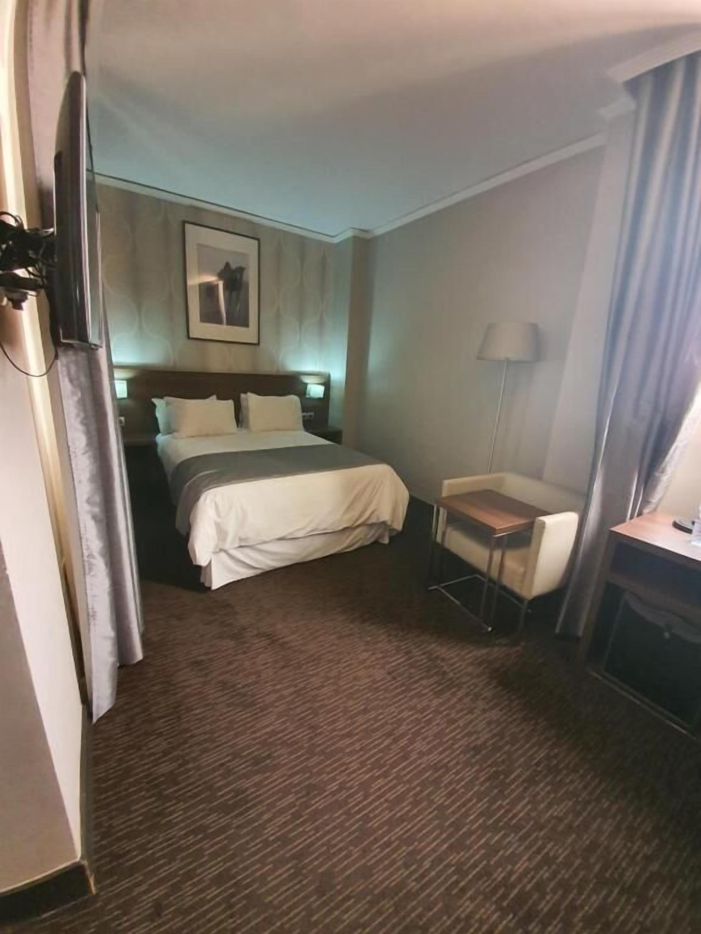 Une chambre d’hôtel avec un lit, un bureau, une chaise, une lampe et un tableau au mur.