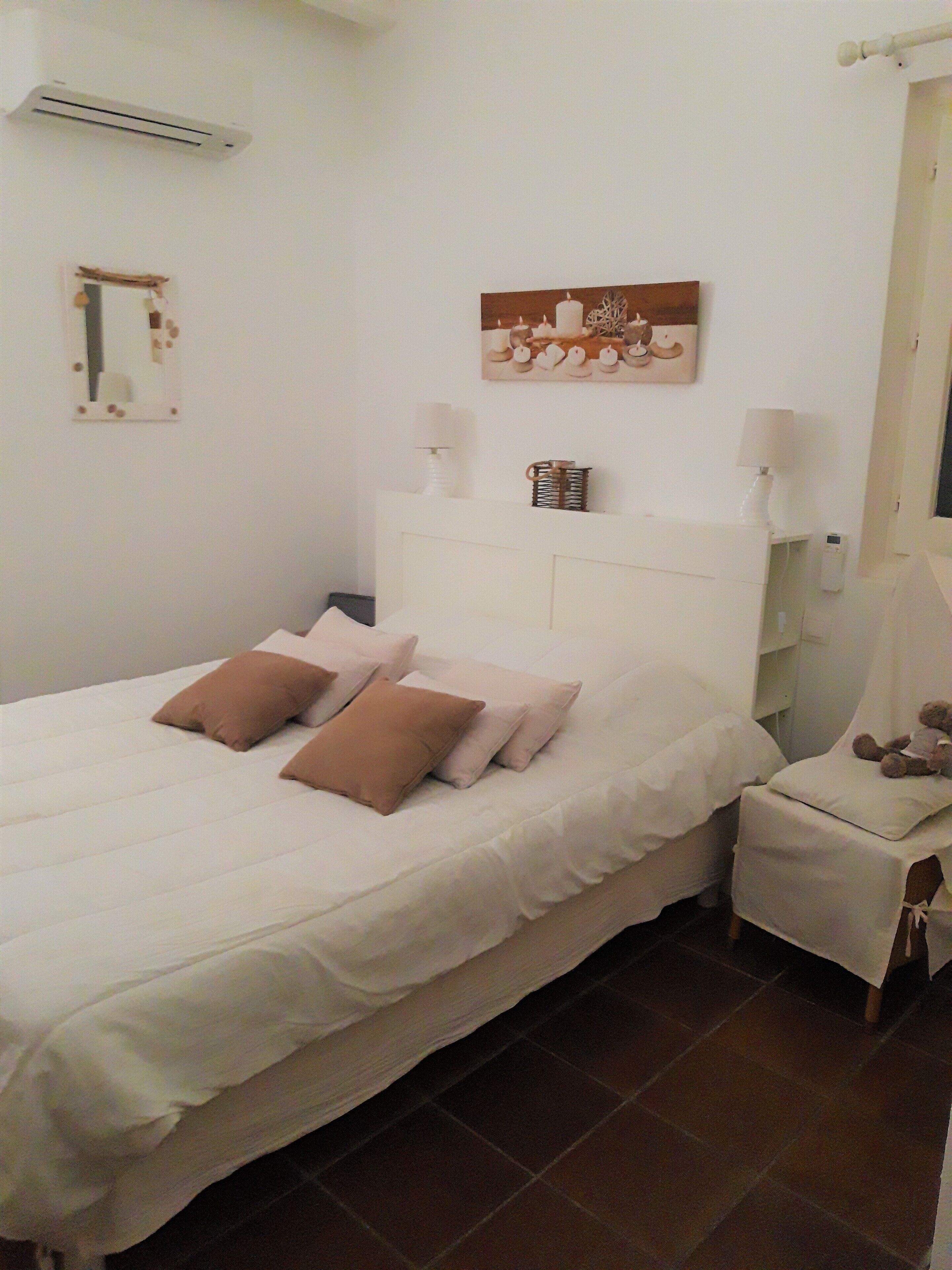 Ein ordentlich bezogenes Bett mit weißen und braunen Kissen, ein Nachttisch mit Lampe und ein kleiner Stuhl mit einem Teddybär.