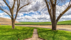 Un sentier traverse une pelouse bien entretenue, bordée de chaque côté de grands arbres et d’une clôture en métal, et offre une vue lointaine sur un champ.