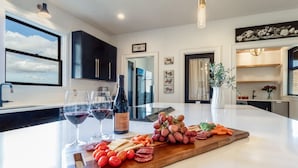 Une cuisine moderne dotée d’un grand îlot central, de verres à vin, d’une bouteille de vin et d’une planche de charcuterie garnie de différents fromages, fruits et légumes.