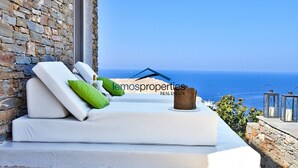A white outdoor seating area with cushions and a wicker basket, overlooking a beach and the sea. - Luxurious villa with a swimming pool, sea and sunset view (Kéa)